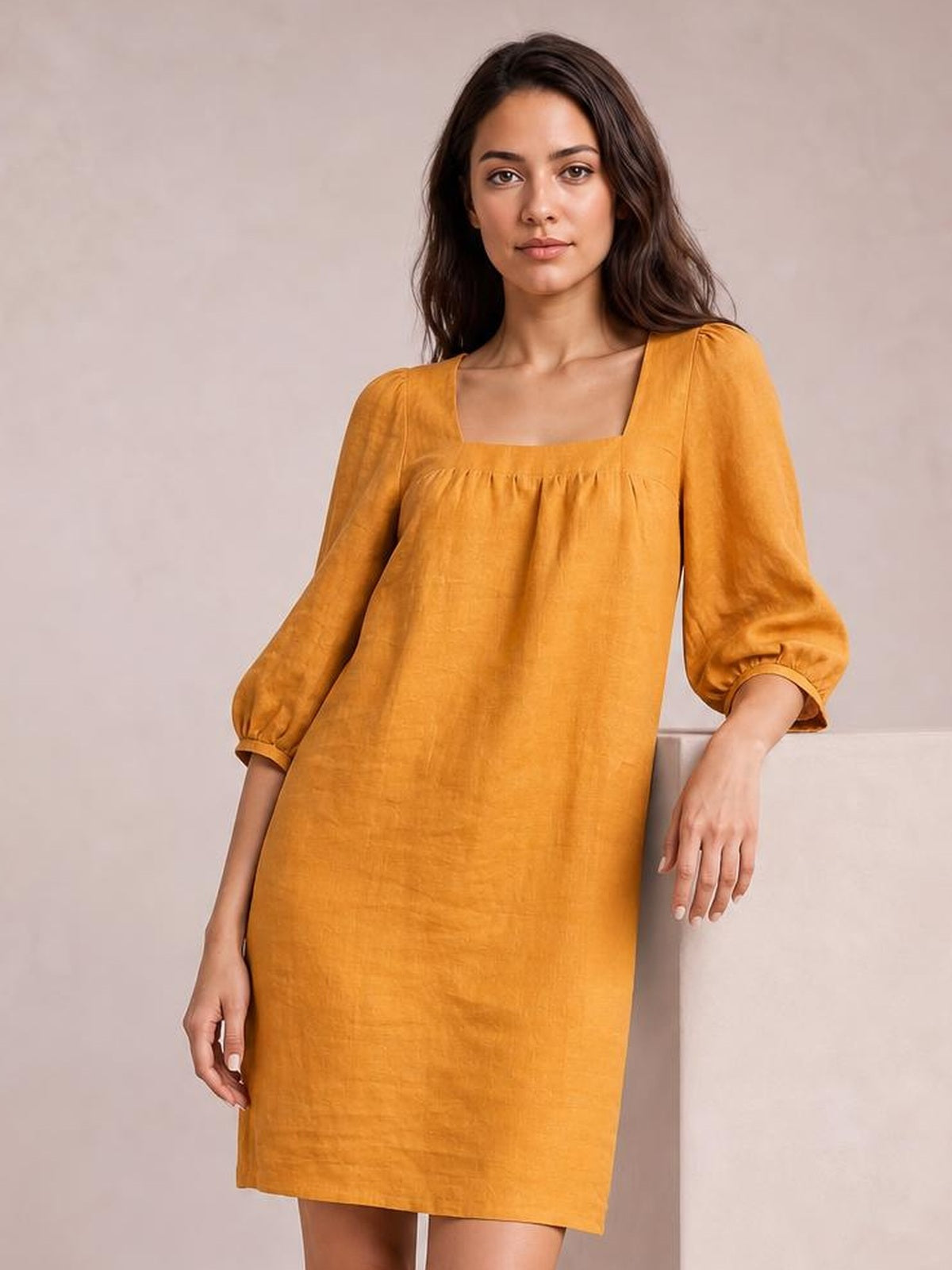 A model leans against a wall wearing a mustard yellow square-neck mini dress with gathered seam detail, puff sleeves, and a relaxed straight silhouette.