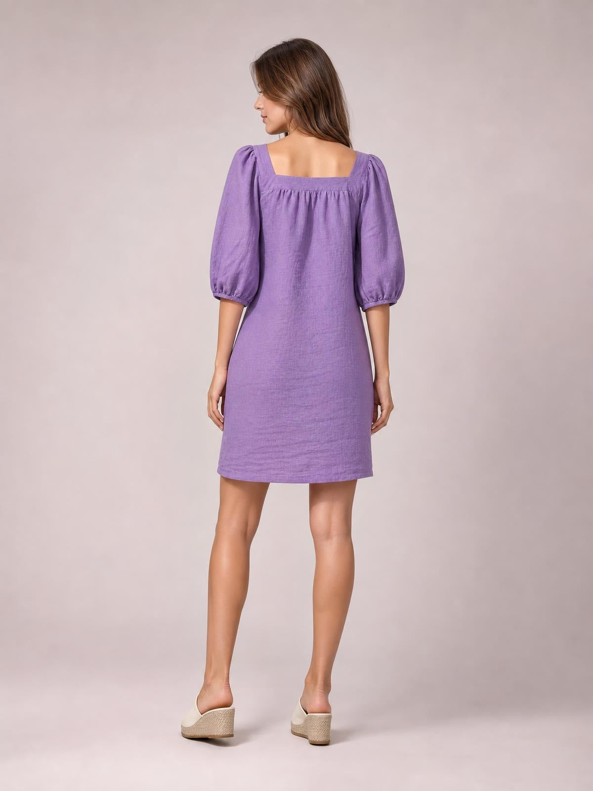 A model stands facing away in a purple square-neck mini dress with puff sleeves, visible seam detail, and a relaxed straight silhouette.