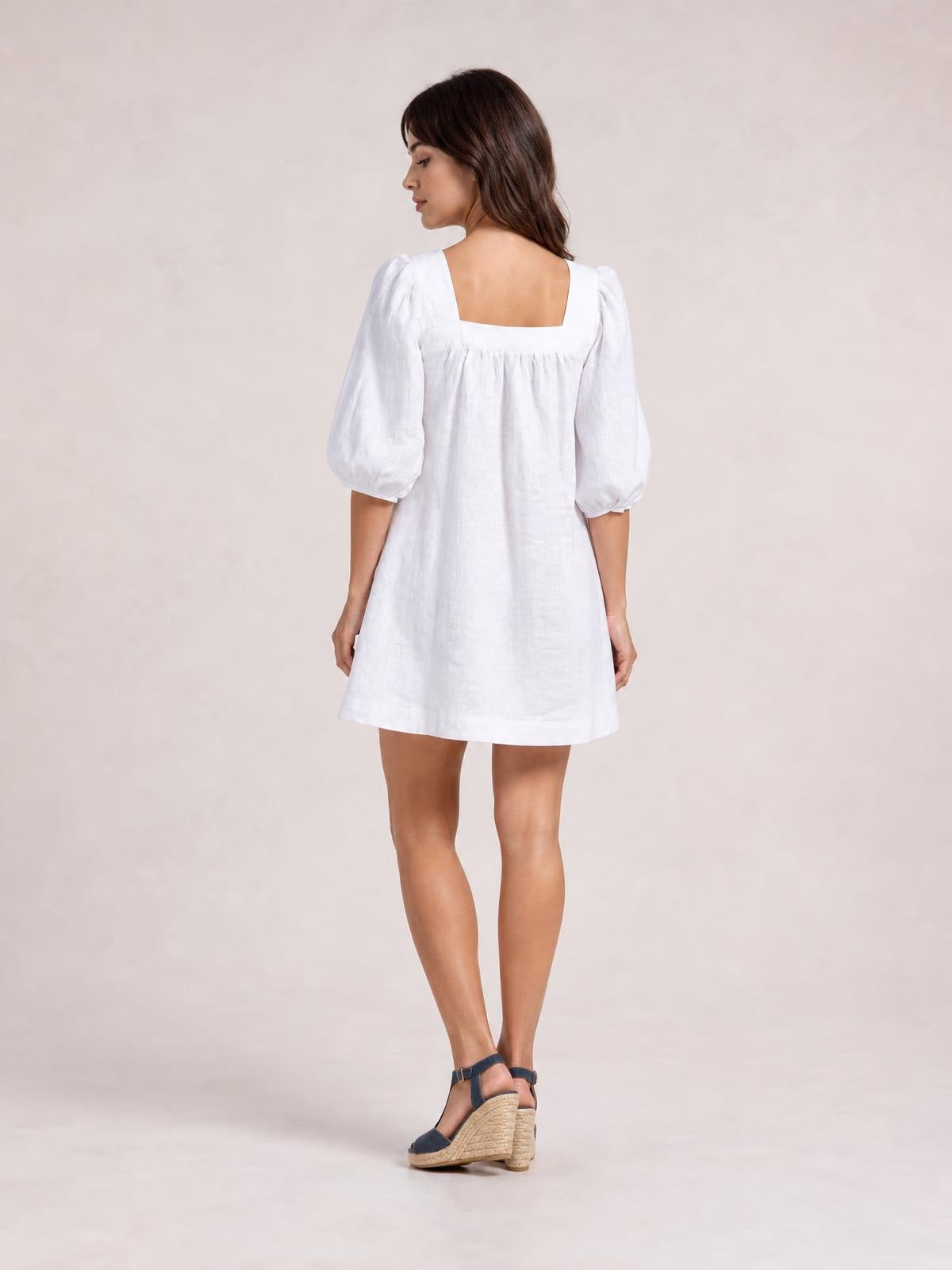 Model turns away in a white square-neck mini dress with puff sleeves, gathered seam detail, and a relaxed, loose A-line silhouette.