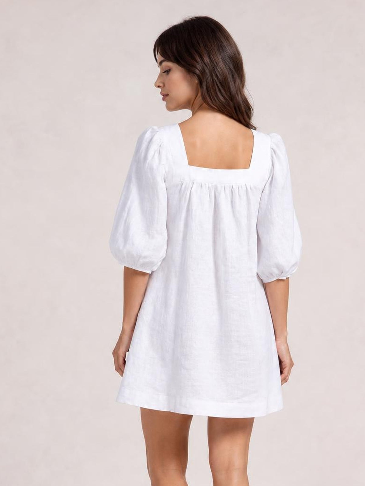 Model shown from the back in a white square-neck puff sleeve mini dress with a relaxed silhouette, gathered seam detail, and three-quarter sleeves.