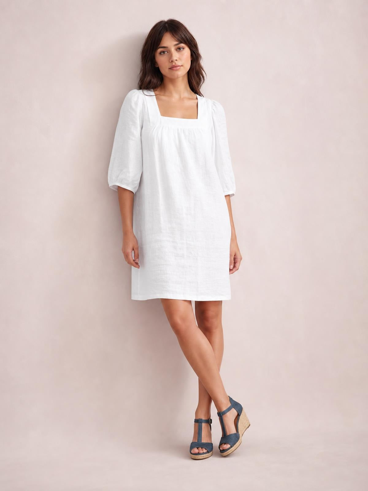 Model poses with crossed legs in a white square-neck mini dress featuring puff sleeves, a relaxed straight silhouette, and visible seam detailing.