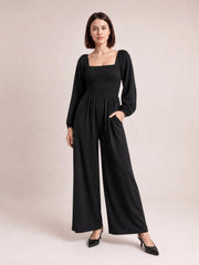 Model stands in a black square-neck jumpsuit with a smocked bodice, long raglan sleeves, wide-leg silhouette, and a practical side pocket.