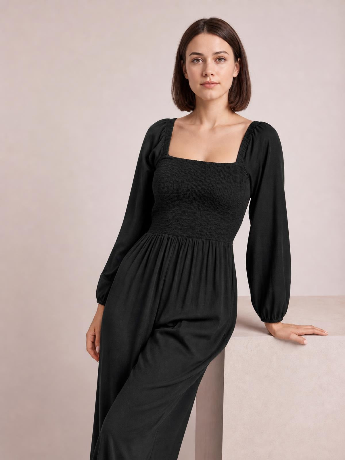 A model leans beside a ledge wearing a solid black jumpsuit with a square neckline, smocked bodice, long raglan sleeves, and a flowing wide-leg silhouette.