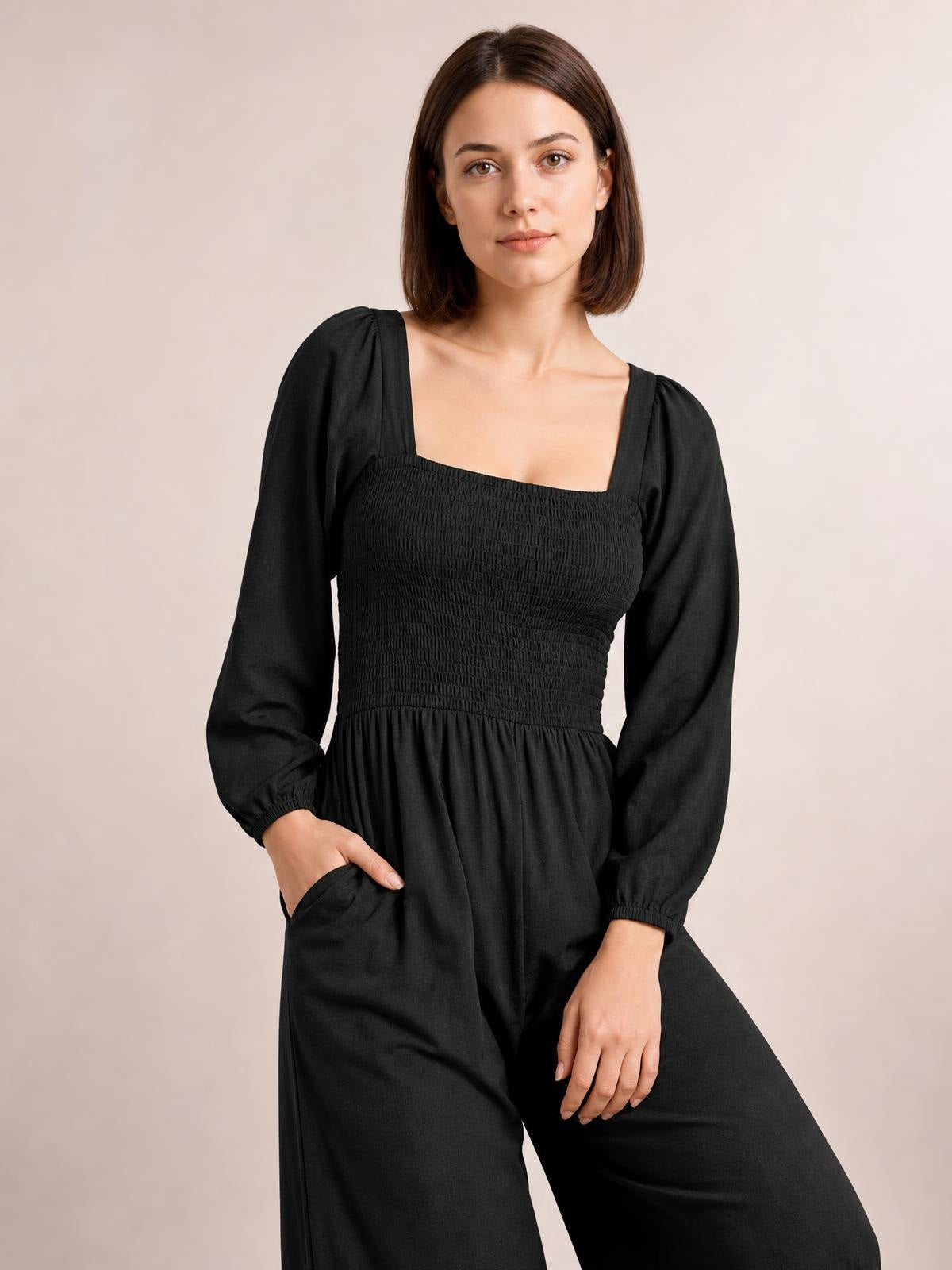 Model wears a black square-neck raglan sleeve jumpsuit with a smocked bodice, long sleeves, wide-leg silhouette, and practical pockets.