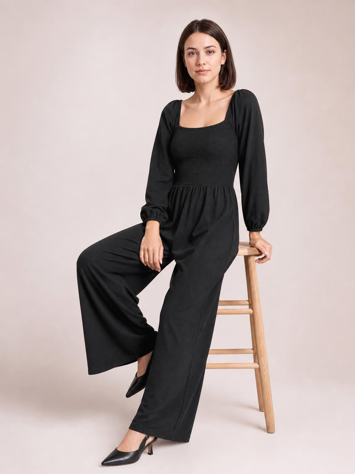Model seated on a stool wears a black long-sleeve raglan jumpsuit with a square neckline, smocked bodice, gathered waist, and wide-leg silhouette.