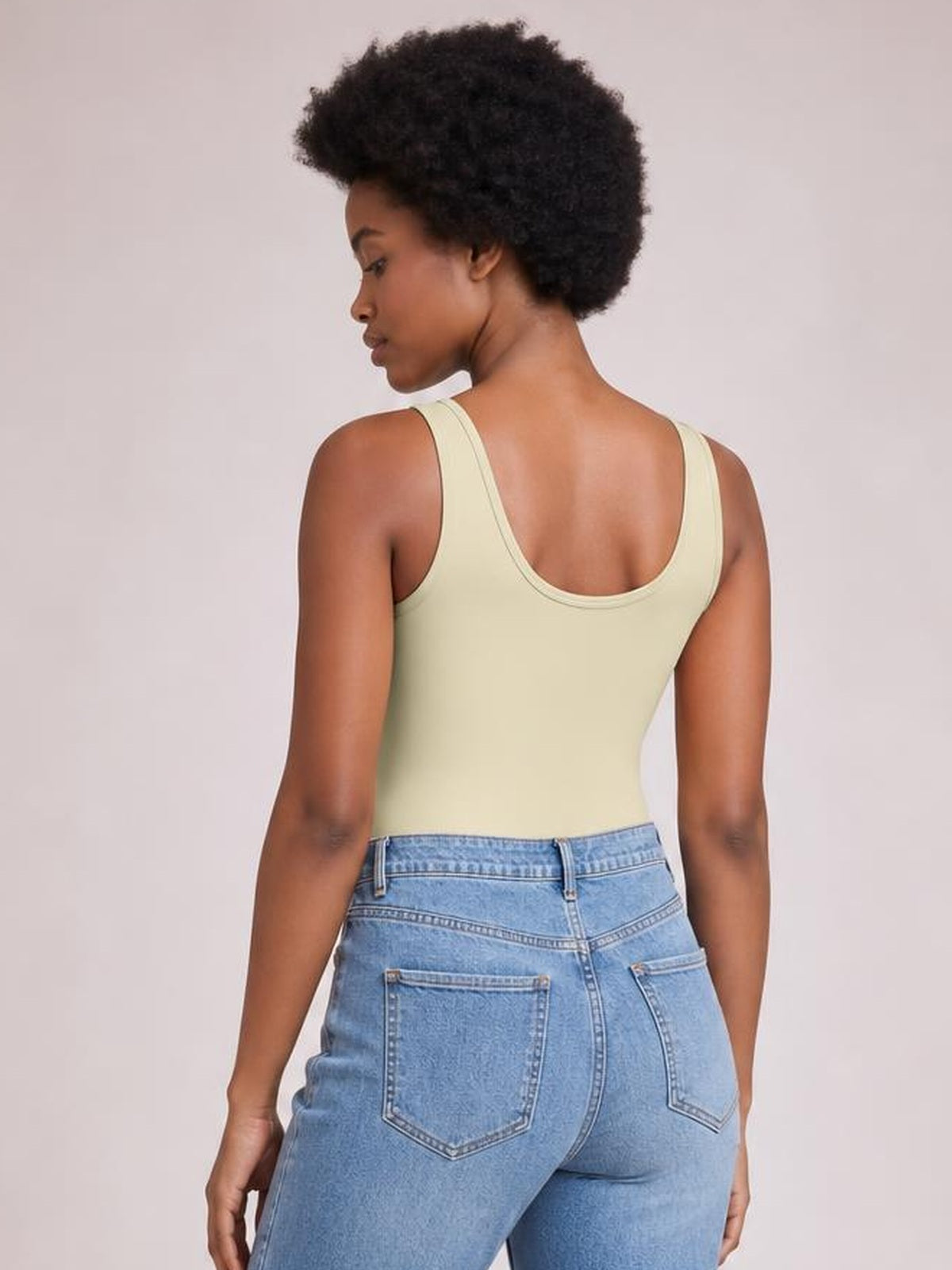 A model shown from the back wears a pale yellow sleeveless bodysuit with wide straps, a low rounded back neckline, and a smooth fitted silhouette.