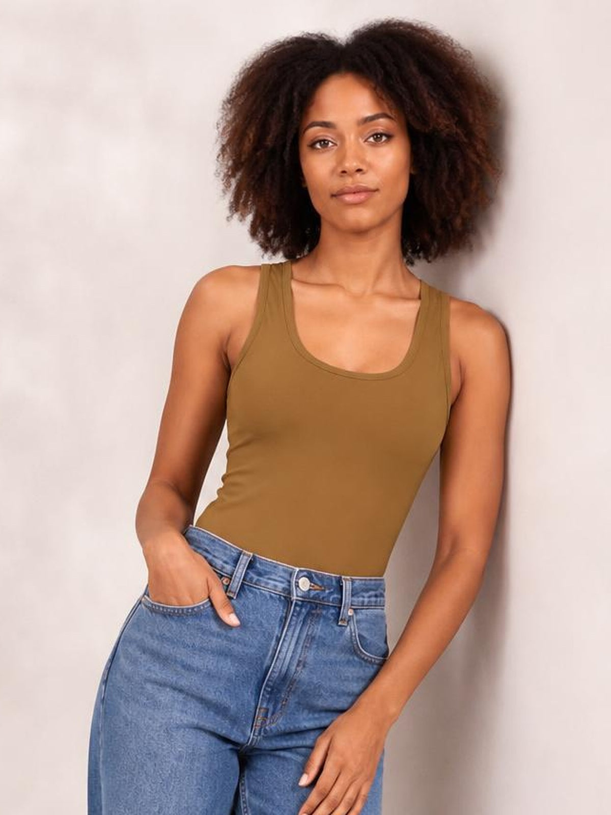 Model poses in a brown sleeveless bodysuit with a rounded square neckline and fitted silhouette, paired with high-rise blue jeans.