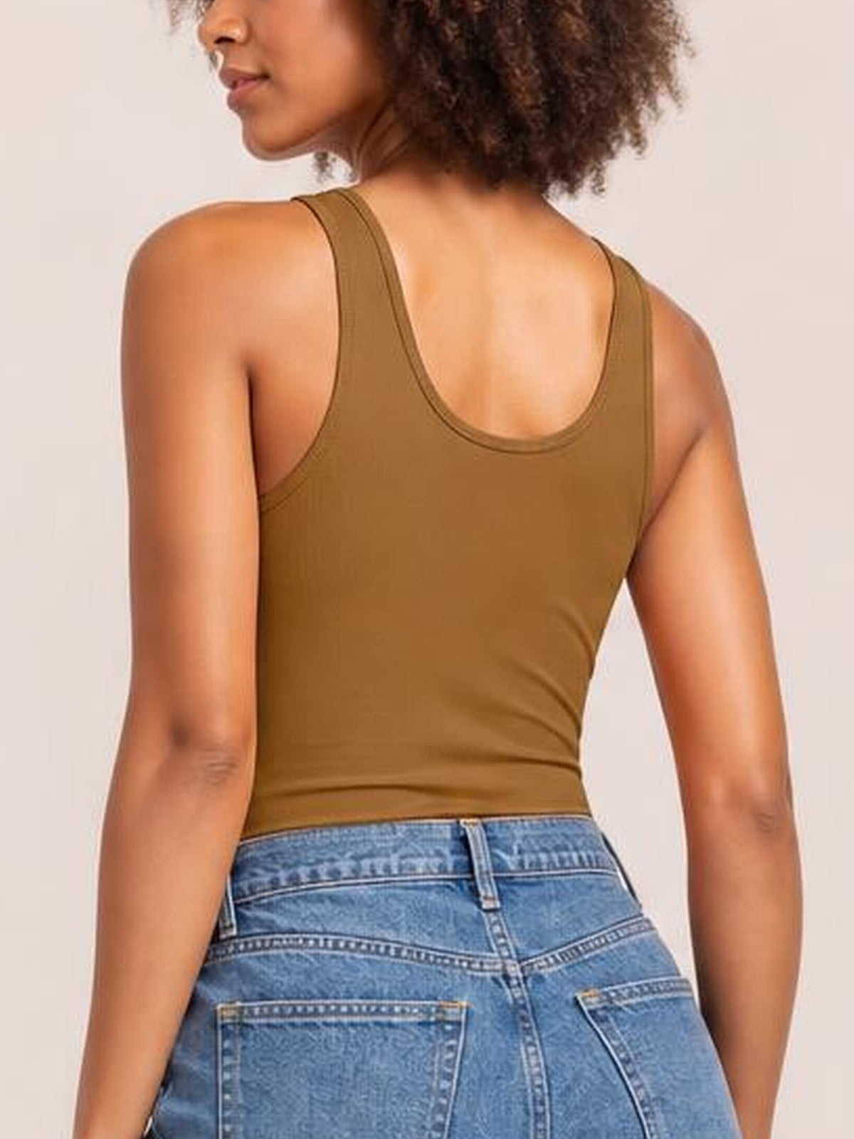 A model turned away wears an olive-brown sleeveless bodysuit with wide straps, a scoop back, and a fitted silhouette tucked into blue jeans.