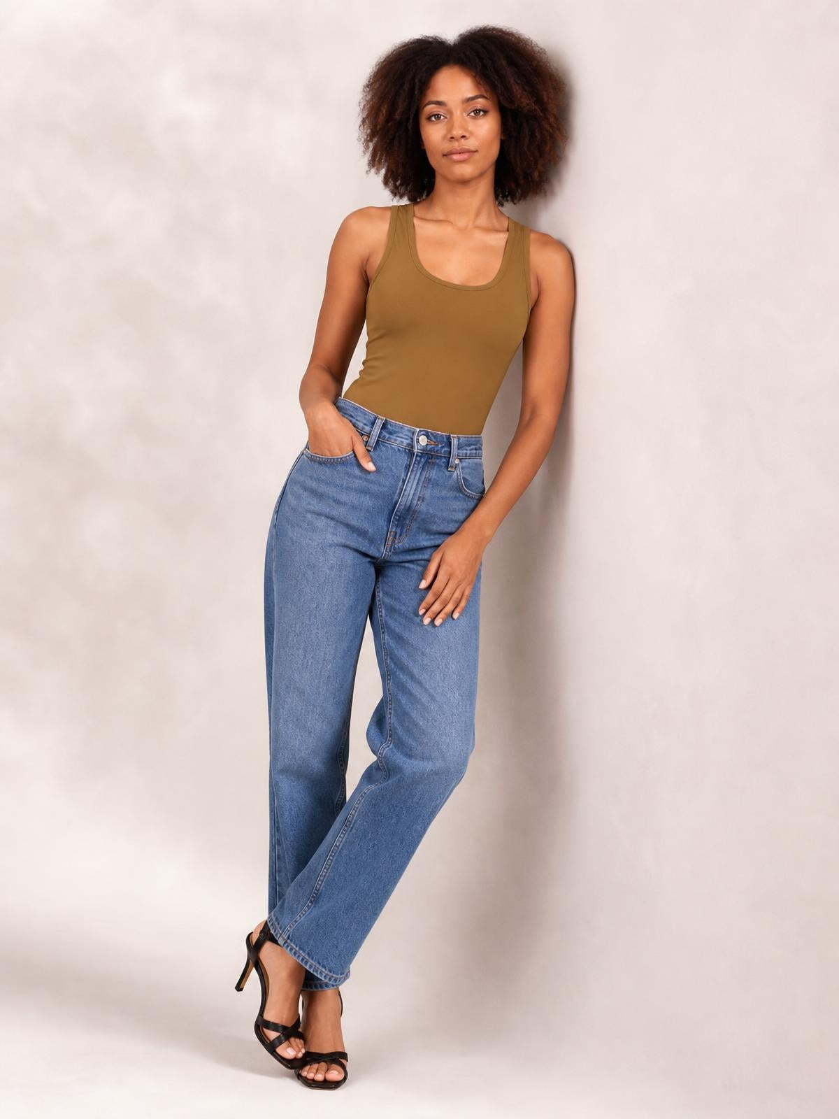Model wears a brown sleeveless bodysuit with wide straps and a fitted silhouette, posing with crossed legs and one hand in her jeans pocket.