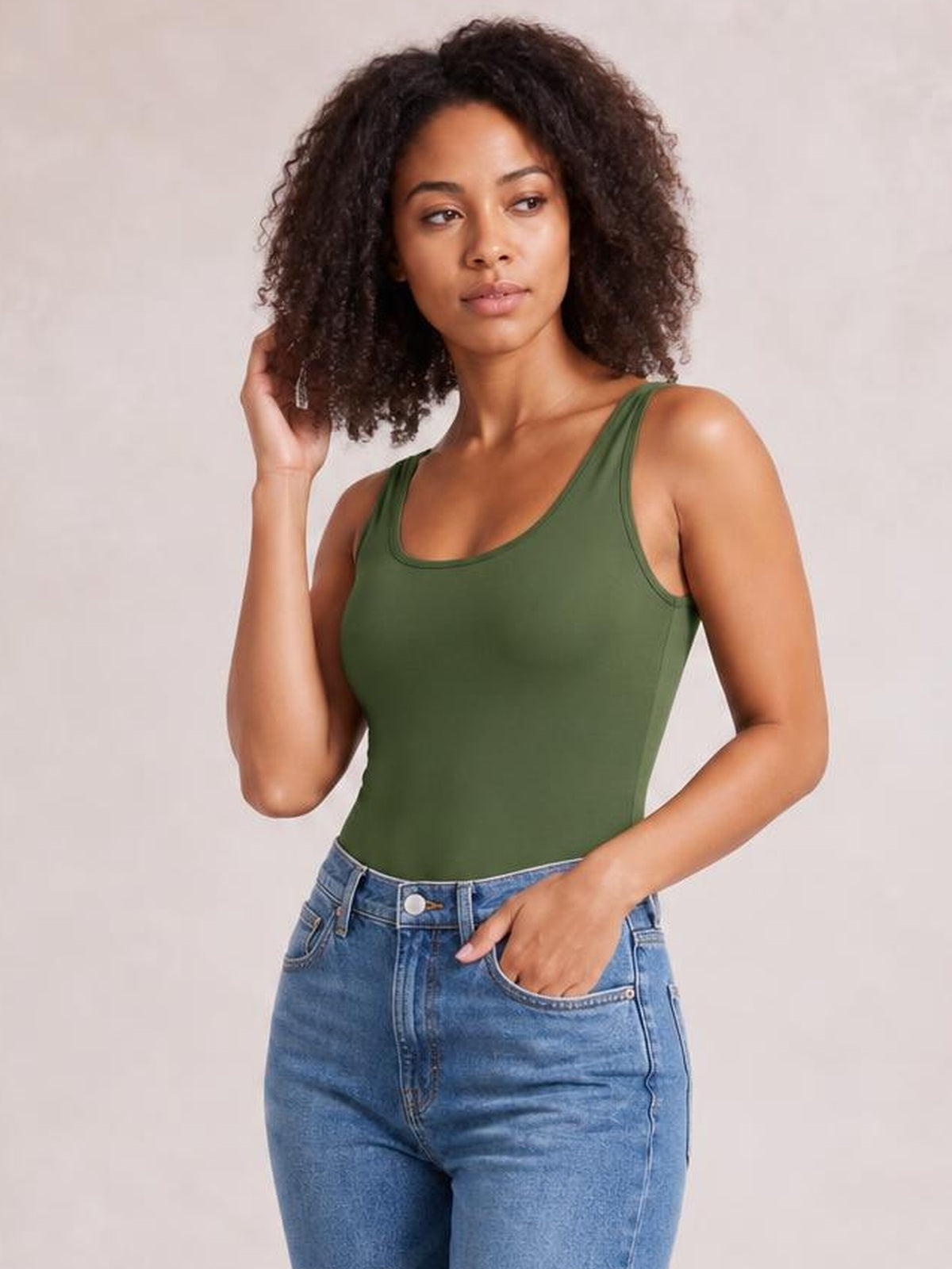 Model wears an opaque olive green sleeveless bodysuit with a fitted silhouette and bound neckline and armholes, posing with one hand in her hair and one in her pocket.