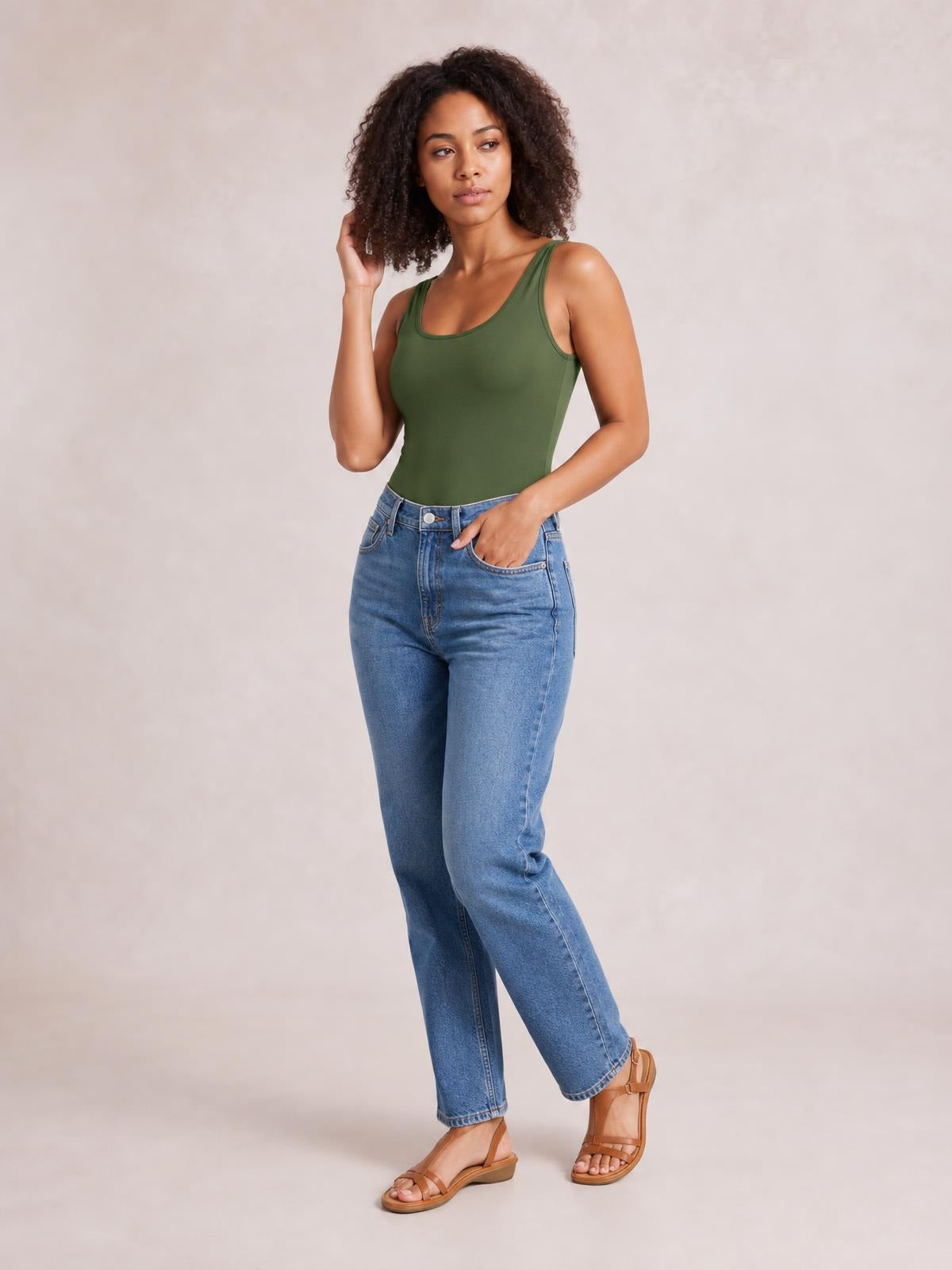 Model wears a fitted olive green square-neck sleeveless bodysuit with wide straps and a smooth, streamlined silhouette, standing with one hand in her jeans pocket.