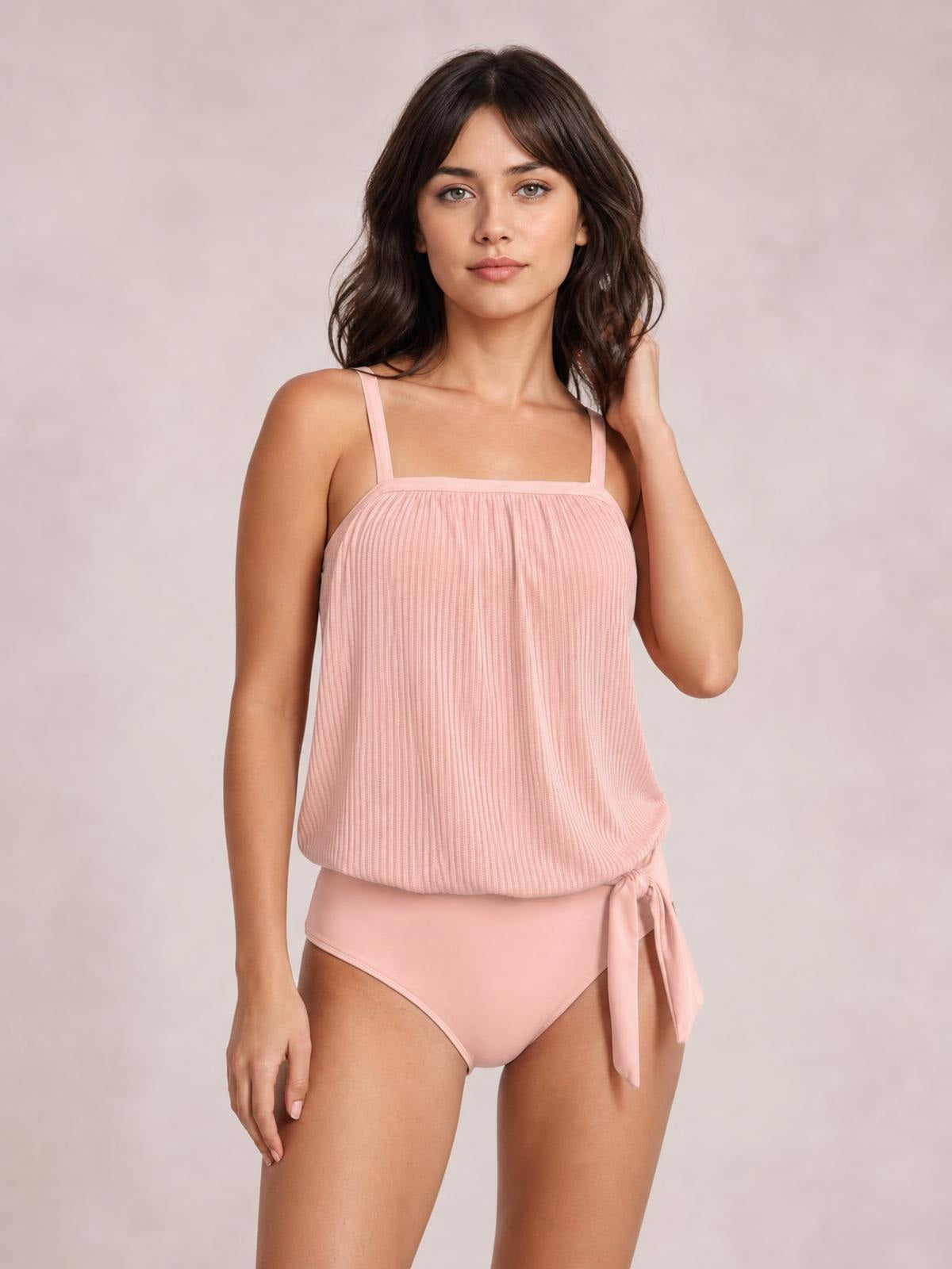 A model poses in a blush pink square-neck spaghetti strap tankini set with a softly gathered ribbed top, relaxed silhouette, and tied side briefs.