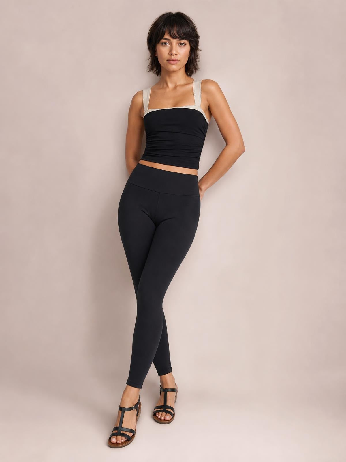 A model poses in a black square-neck cami with wide contrasting straps, a fitted cropped silhouette, and subtle ruching, paired with black leggings.
