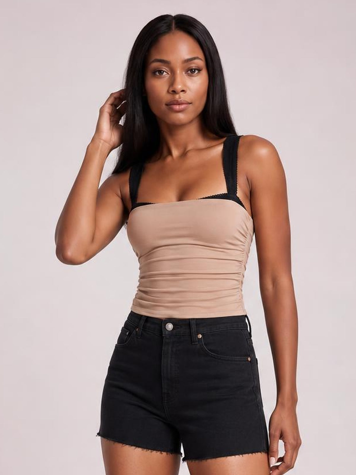 A model poses in a beige square neck cami with wide straps, a fitted ruched silhouette, and subtle stretch, paired with black denim shorts.