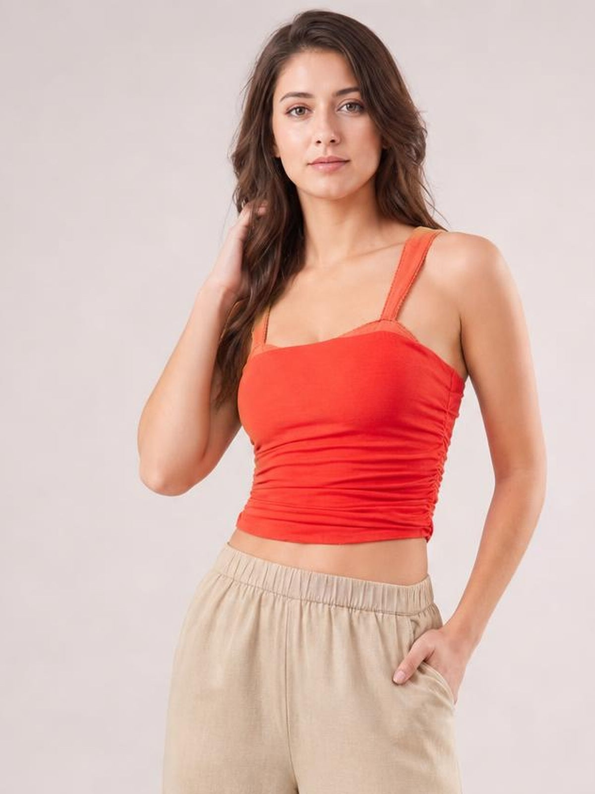 A model poses in a red square-neck wide-strap cami with a fitted, ruched silhouette, lifting one hand to her hair and resting the other in a pocket.