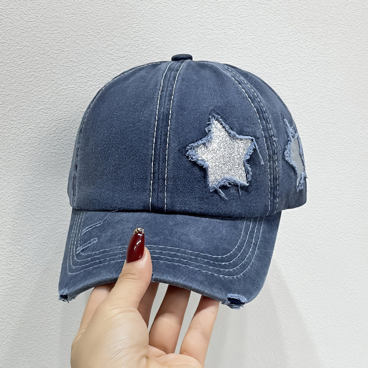 A hand holds a navy adjustable baseball cap with a softly structured six-panel crown, curved brim, contrast seams, distressed edges, and star appliqués.