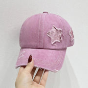 A hand holds a pink adjustable baseball cap featuring a softly structured six-panel crown, curved brim, distressed edging, and a star appliqué.