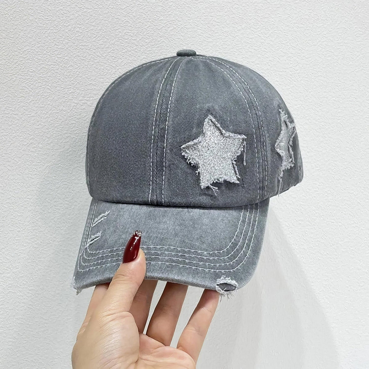 A gray adjustable baseball cap with a softly structured six-panel crown, curved brim, contrast seams, star appliqué details, and distressed edging is held by hand.