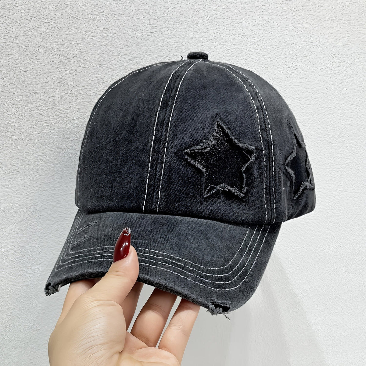 A hand holds a black Star adjustable baseball cap with a six-panel crown, curved brim, contrasting seams, star appliqué, and distressed edging.