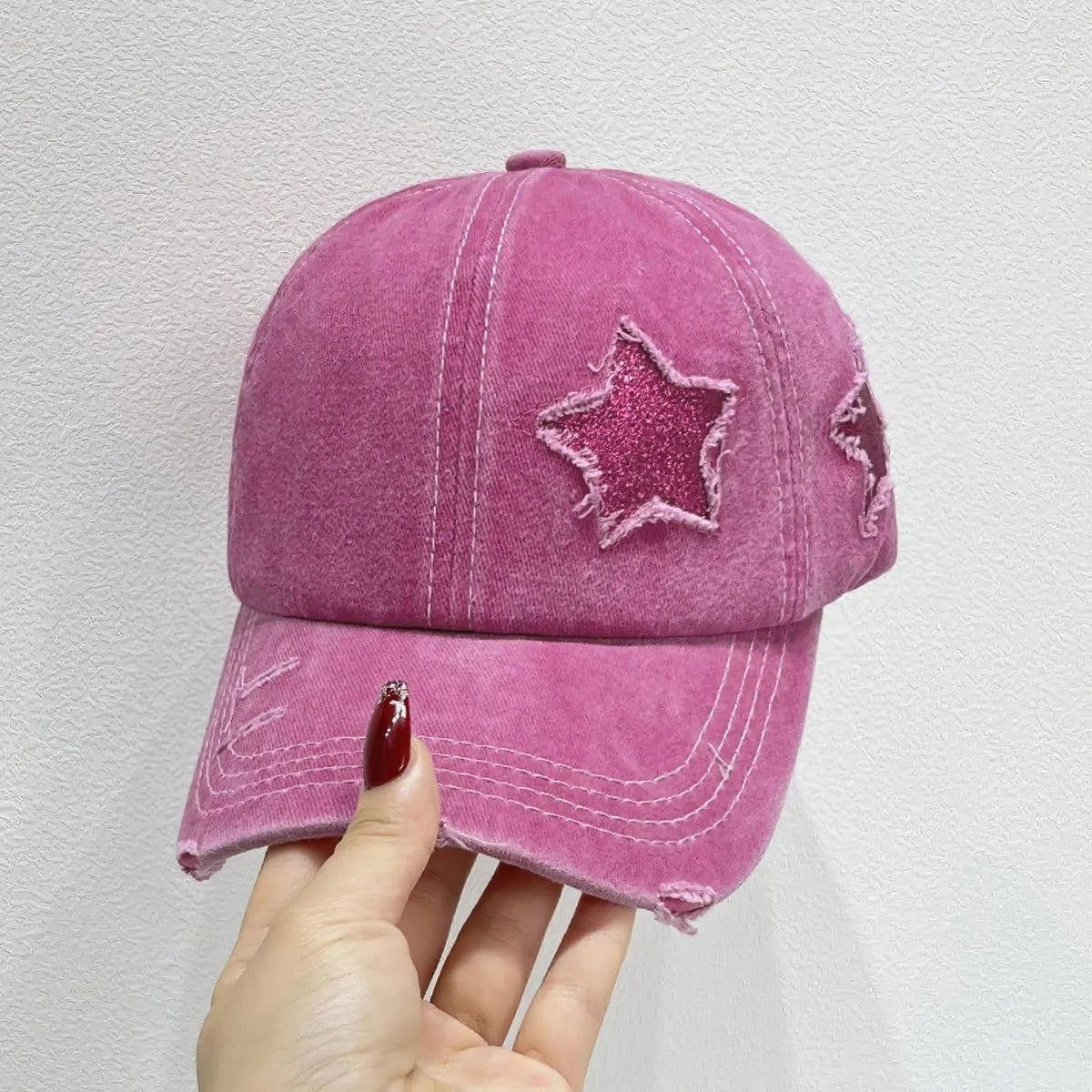 A hand holds a pink cotton adjustable baseball cap with a softly structured six-panel crown, curved brim, star appliqué, and distressed edging.