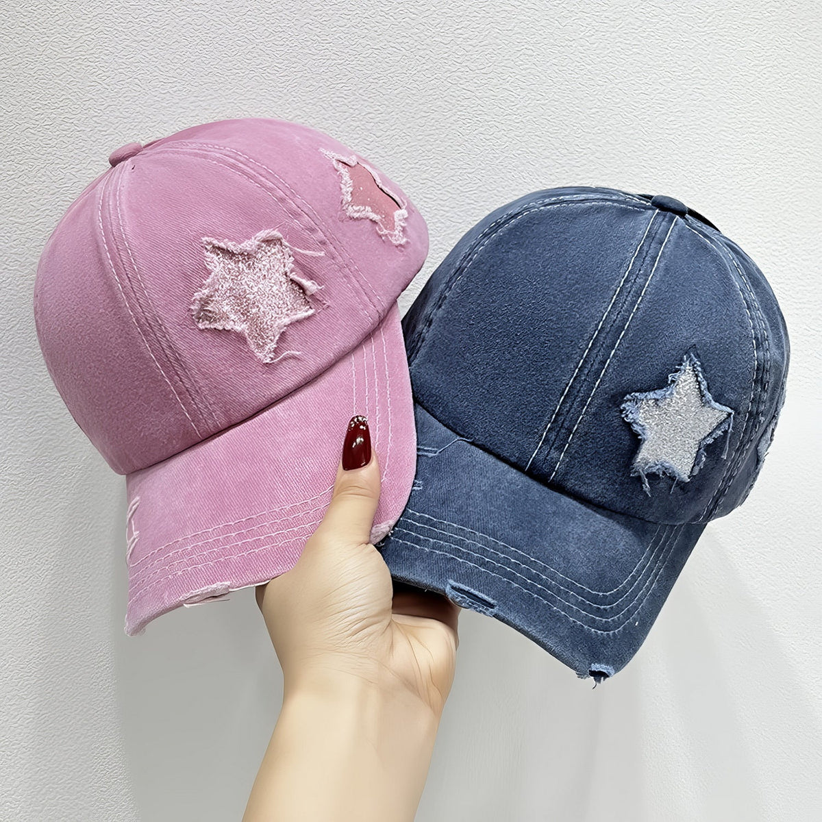 A hand displays pink and navy cotton baseball caps with curved brims, six-panel seams, star appliqué details, and distressed edges.