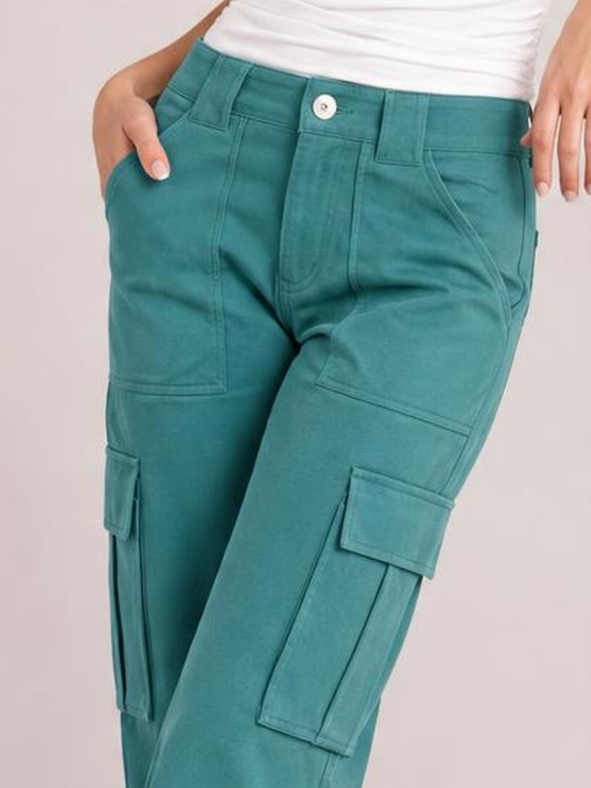 A model wears teal straight-leg cargo pants with multiple flap pockets, belt loops, button closure, and visible contrast stitching, posing with hands in the pockets.