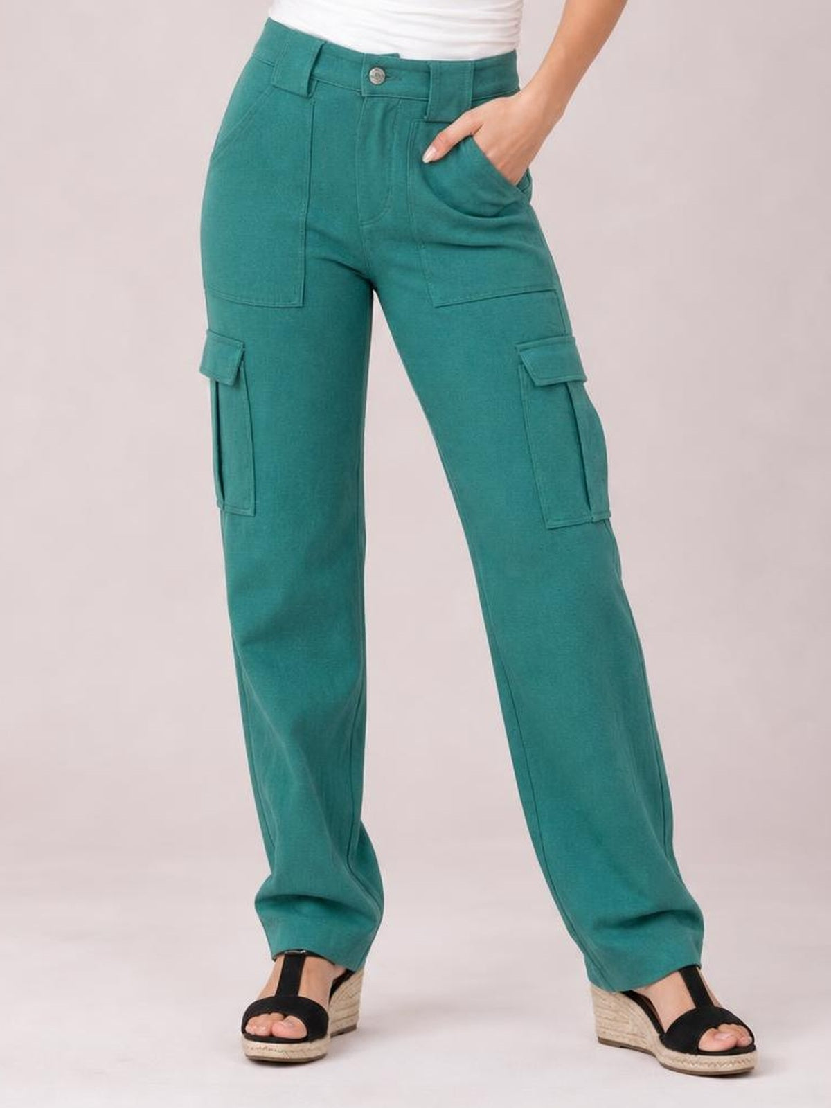 Model wears teal straight-leg cargo pants with a button closure, belt loops, front pockets, and multiple flap cargo pockets, posing with one hand in a pocket.