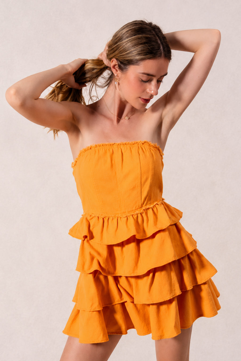 Model poses with her hands in her hair wearing a bright gold strapless dress with a fitted smocked bodice, ruffle waist, and cascading layered skirt.