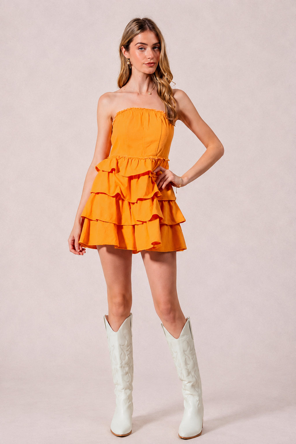 A model poses with one hand on her hip in a bright gold-orange strapless dress with a smocked bodice and cascading ruffle tiers.
