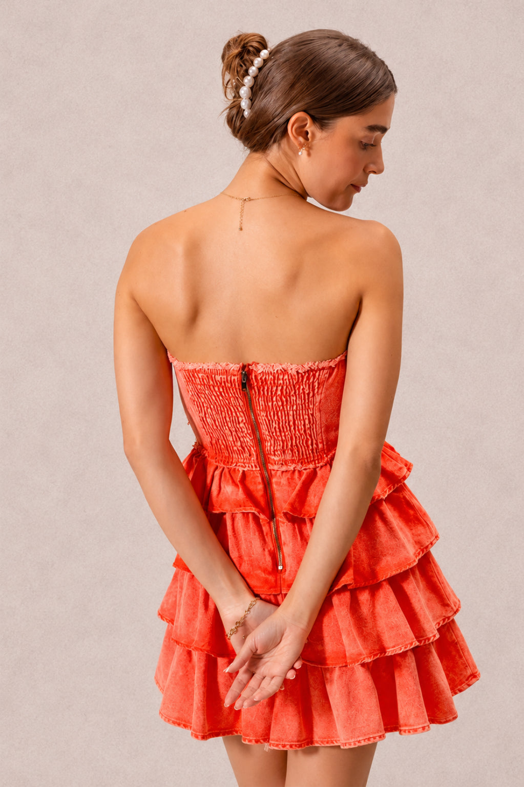 A model poses with her hands behind her in an orange strapless ruffle dress featuring a smocked back, center zipper, and short layered tiered skirt.