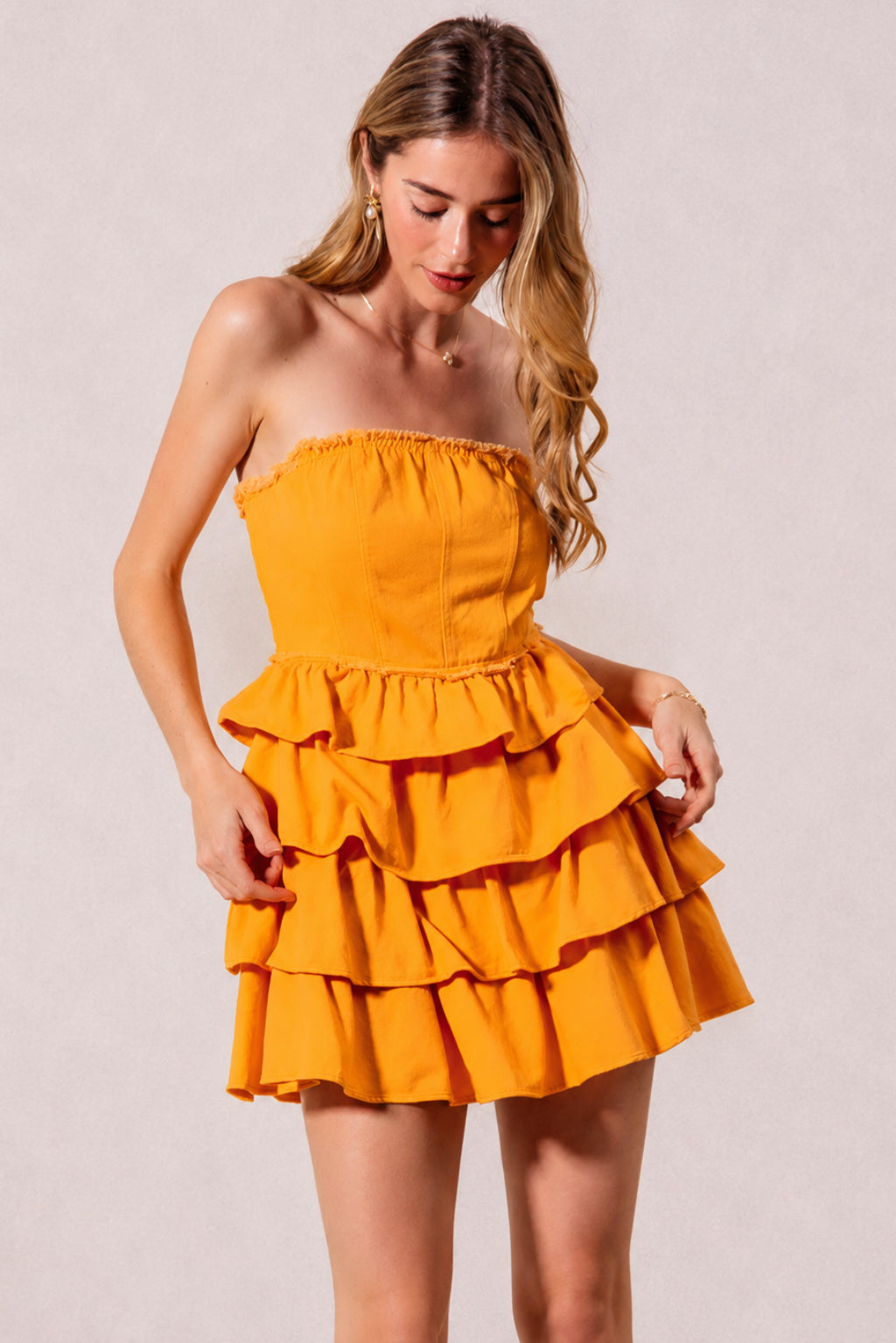 Model poses in a bright gold strapless dress with a fitted smocked bodice, softly gathered neckline, and cascading ruffle tiers creating a playful layered silhouette.
