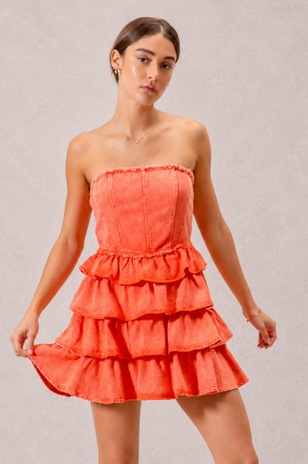 A model poses in an orange strapless dress with a structured bodice, gathered waist, and cascading ruffle layers forming a playful short tiered silhouette.