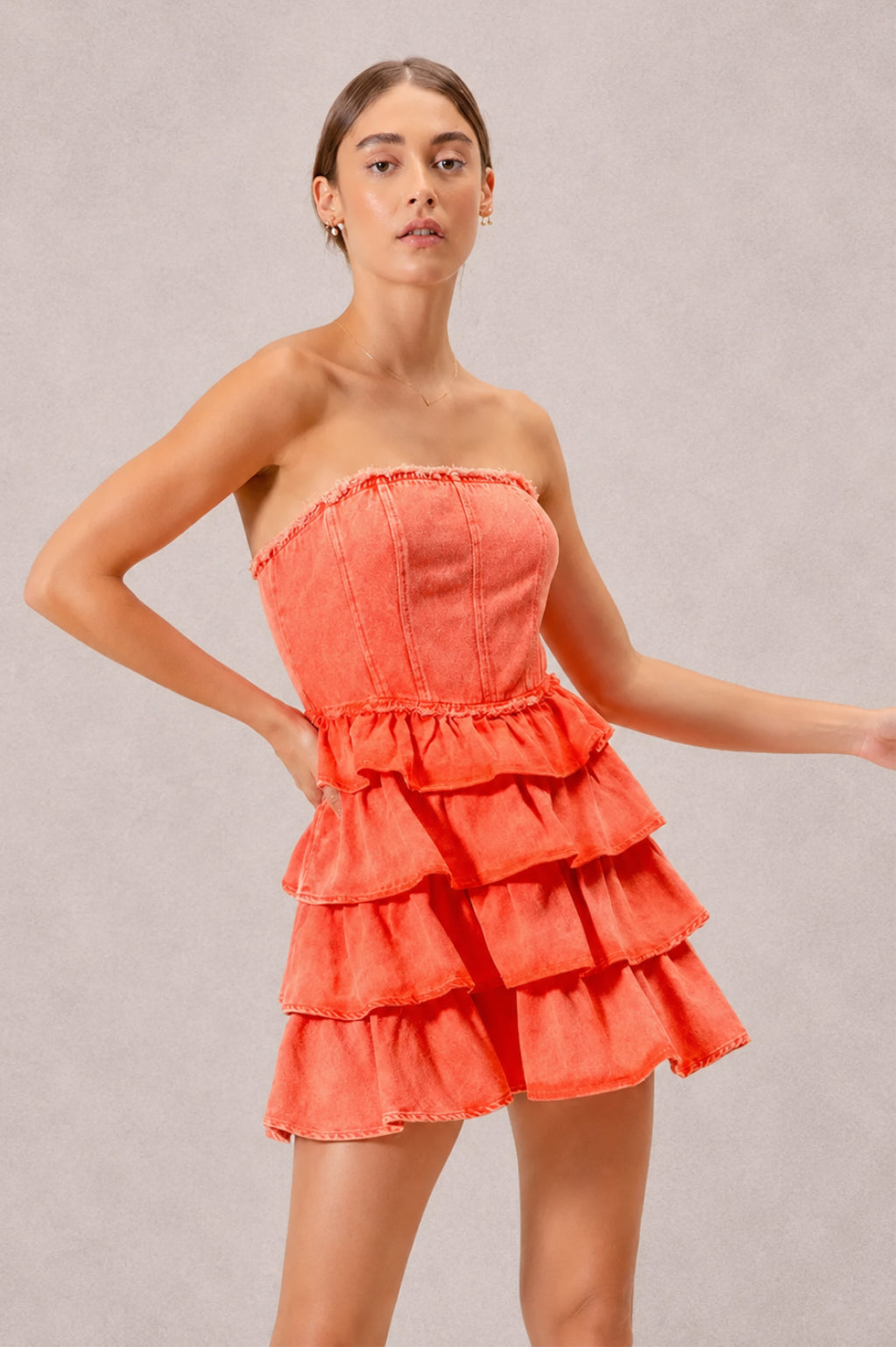 A model poses in an orange strapless dress with a structured bodice, ruffle-trimmed waist, and cascading tiered layers creating a flirty short silhouette.