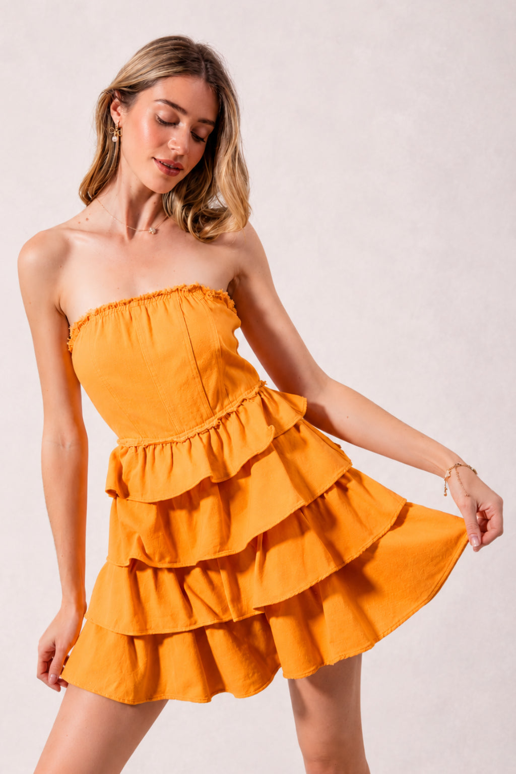 Model poses in a gold strapless dress with a smocked bodice, softly gathered neckline, and cascading layered ruffle tiers forming a flared silhouette.