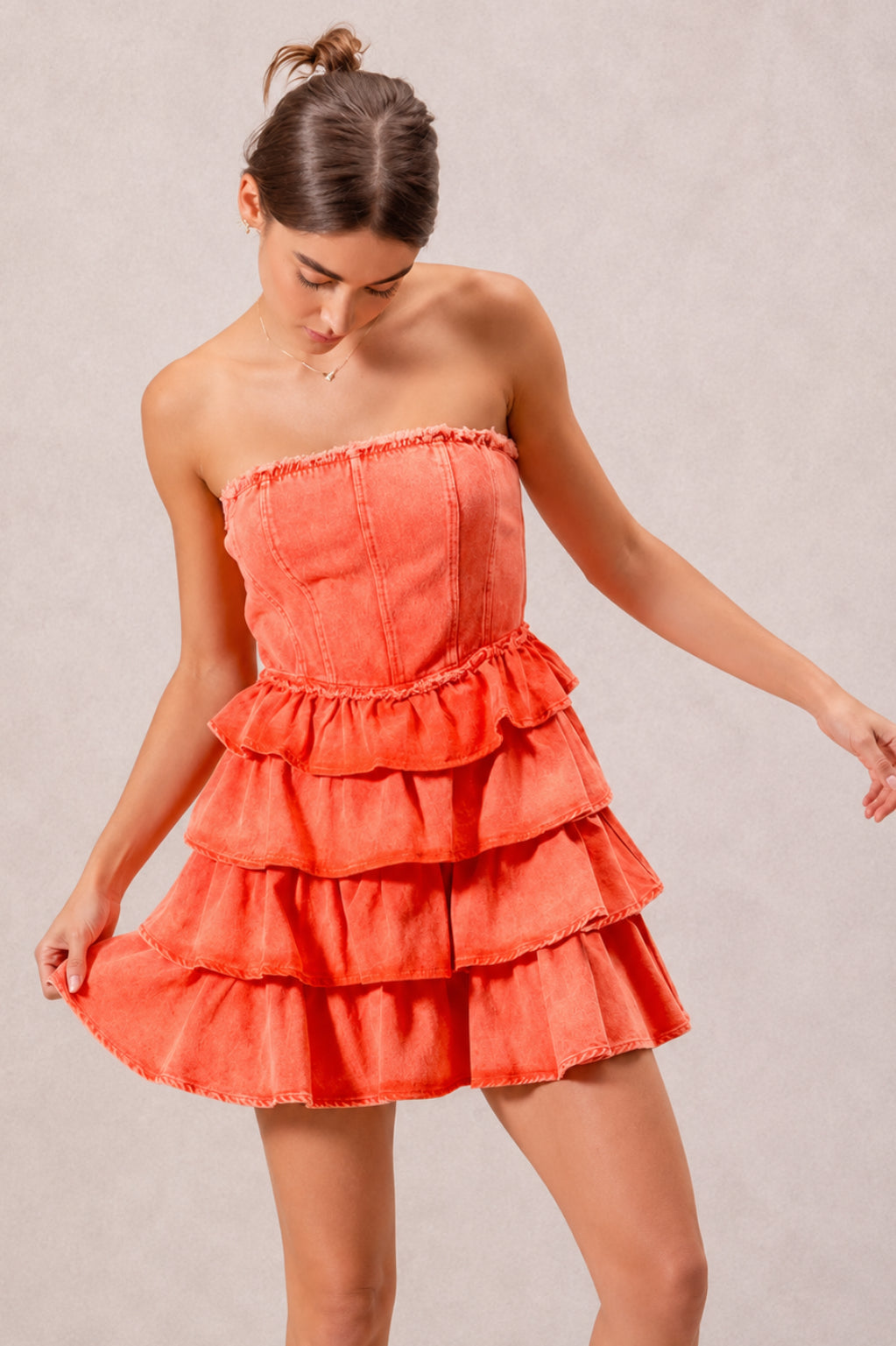 Model poses in a vibrant orange strapless ruffle dress with a structured bodice, smocked waist, and cascading tiered layers forming a playful short silhouette.