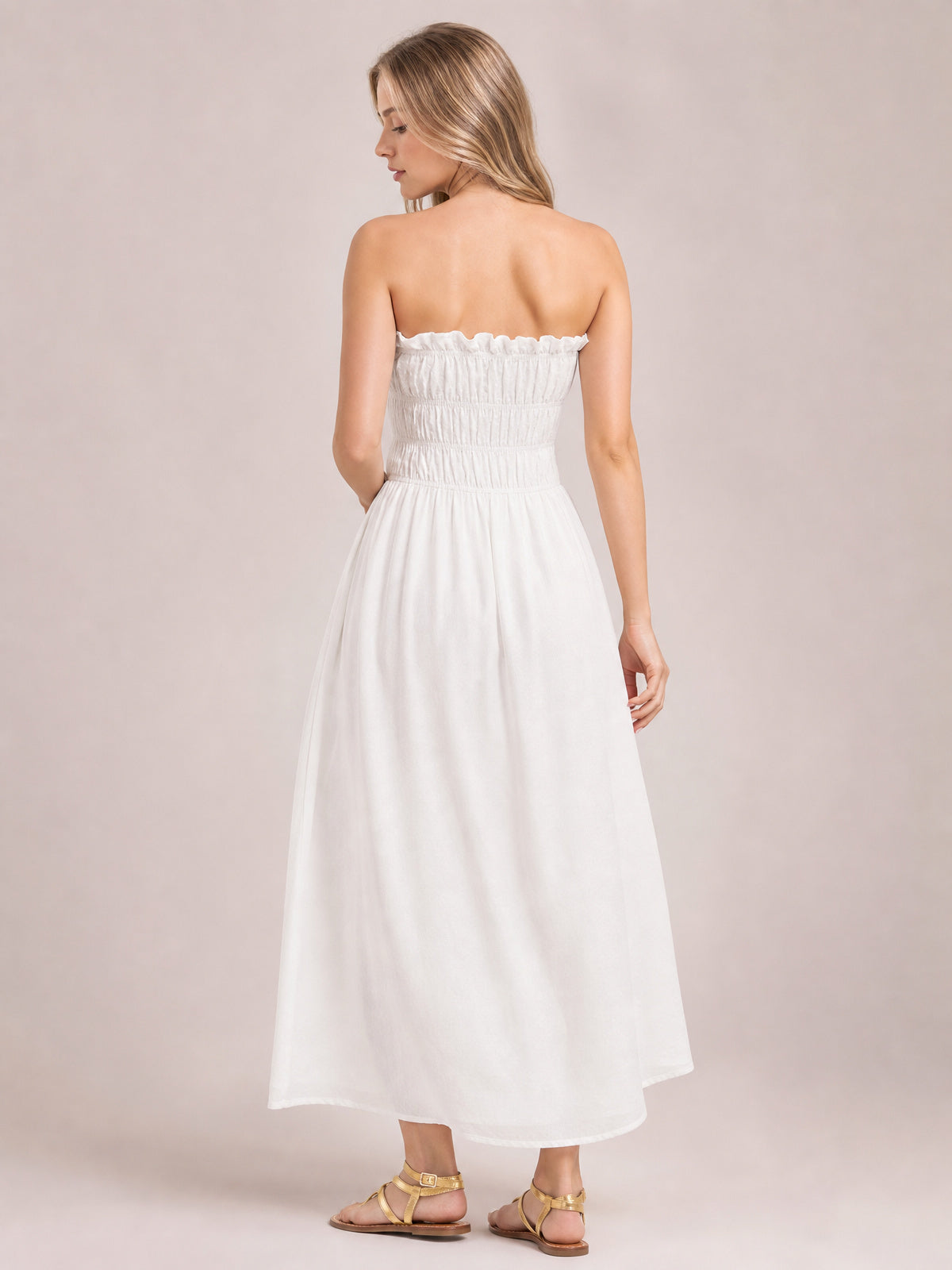 A model looks over her shoulder in a white strapless smocked dress with a frilled neckline, gathered bodice, flowing silhouette, and asymmetric hem.