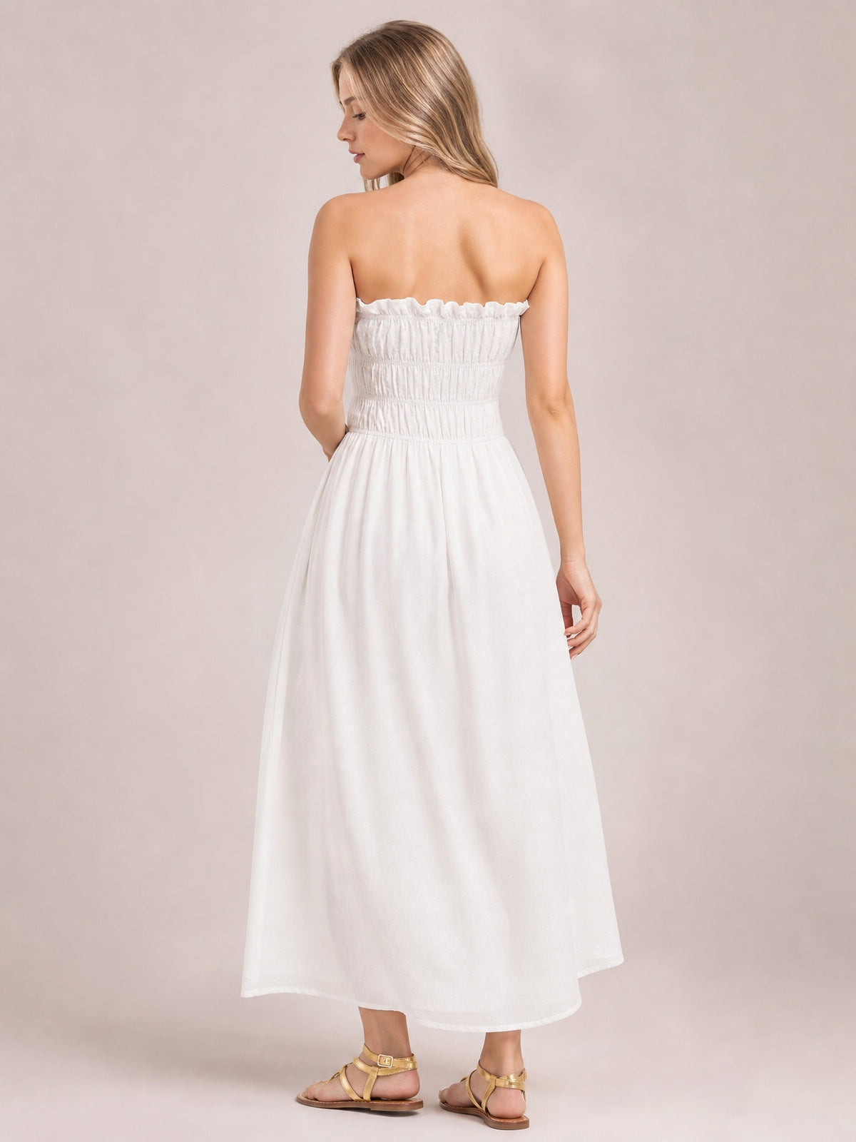 A model looks over her shoulder in a white strapless smocked dress with a frilled neckline, gathered bodice, flowing silhouette, and asymmetric hem.