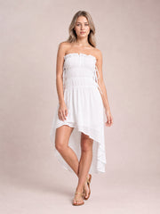 Model poses in a white strapless smocked mini dress with a frilled neckline, side ties, lined body, and graceful asymmetric high-low hem.