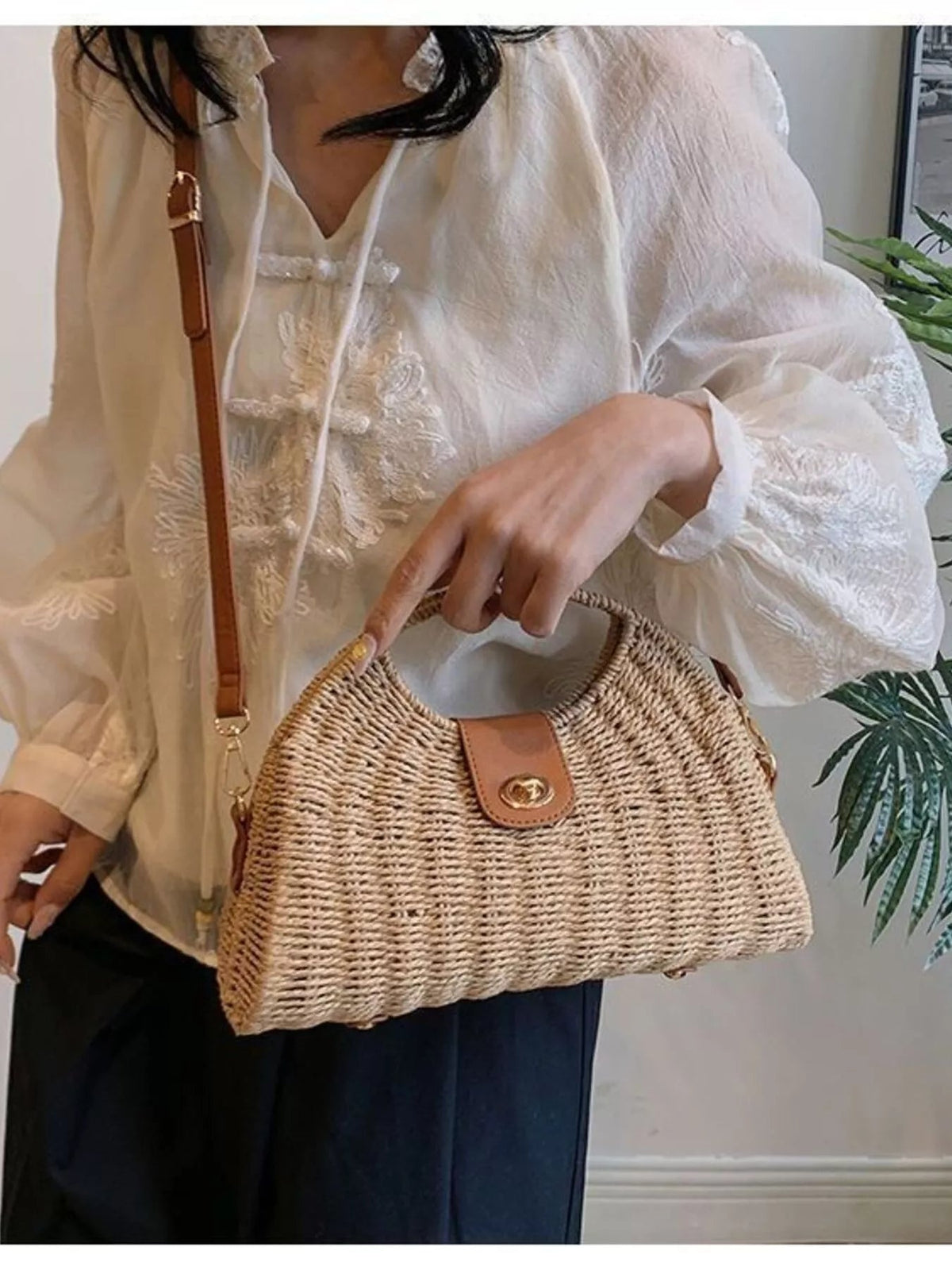 A model holds a medium tan straw woven crossbody bag with an adjustable brown strap, rectangular silhouette, and leather tab closure.