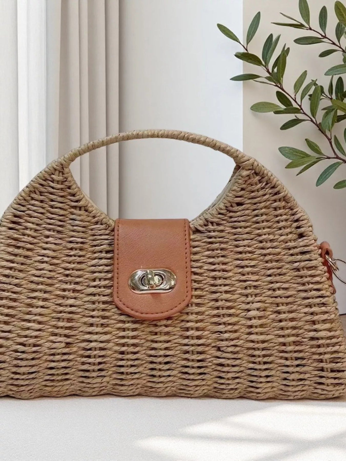 Medium tan straw woven bag with a structured trapezoid silhouette, rounded top handle, brown flap closure, and polished silver clasp.