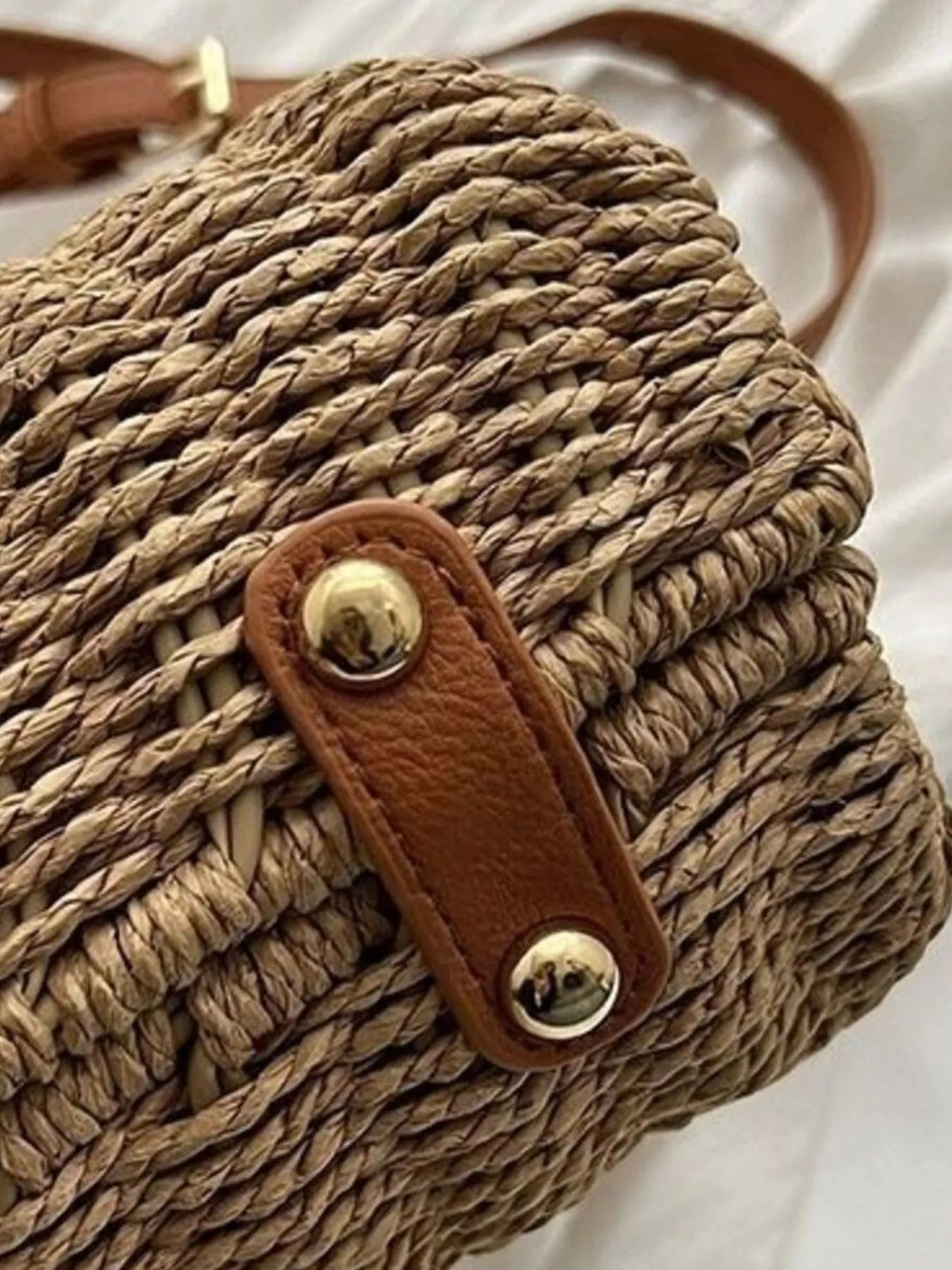 Close-up of a medium natural straw woven crossbody bag with a rounded silhouette and brown leather adjustable strap secured by polished metal studs.