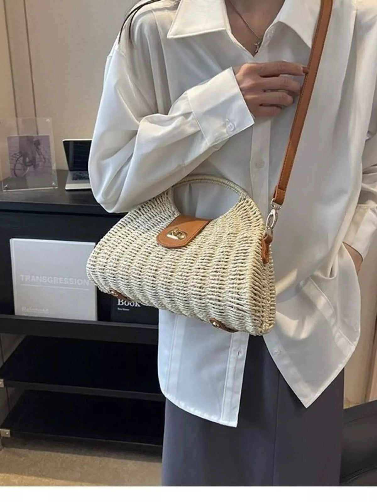 A model wears a medium cream straw woven crossbody bag with an adjustable brown strap, rounded rectangular silhouette, and leather-look flap closure.