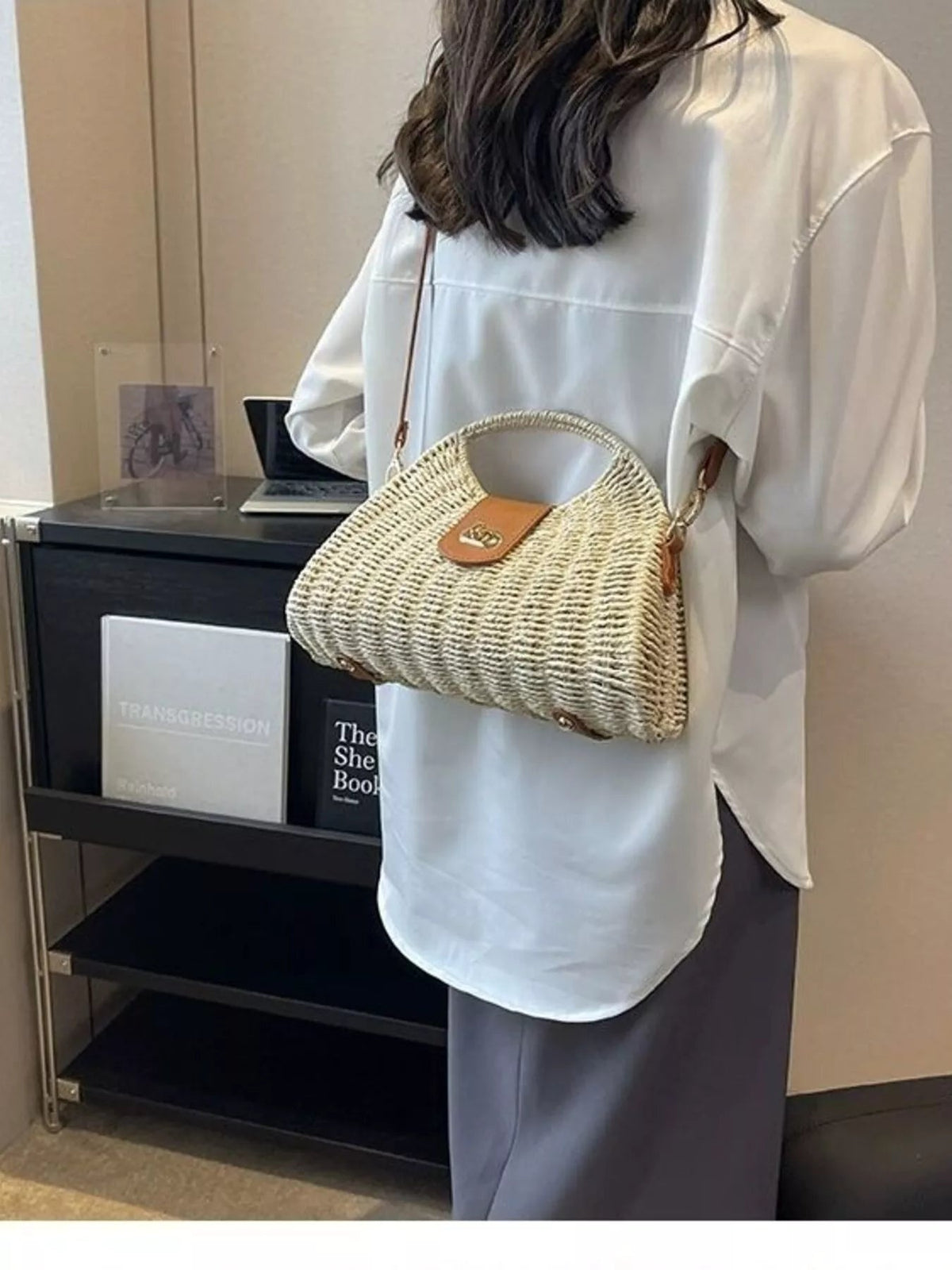A model wears a medium cream straw woven crossbody bag with an adjustable strap, curved top handle, brown flap closure, and rounded silhouette.