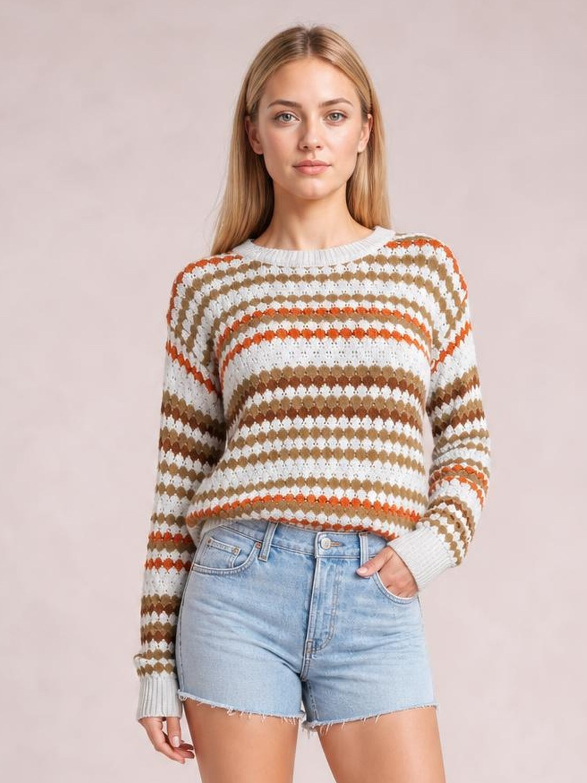 Model wears a cream drop-shoulder sweater with brown and orange striped eyelet pattern, ribbed crew neckline and cuffs, posed with one hand in her shorts pocket.