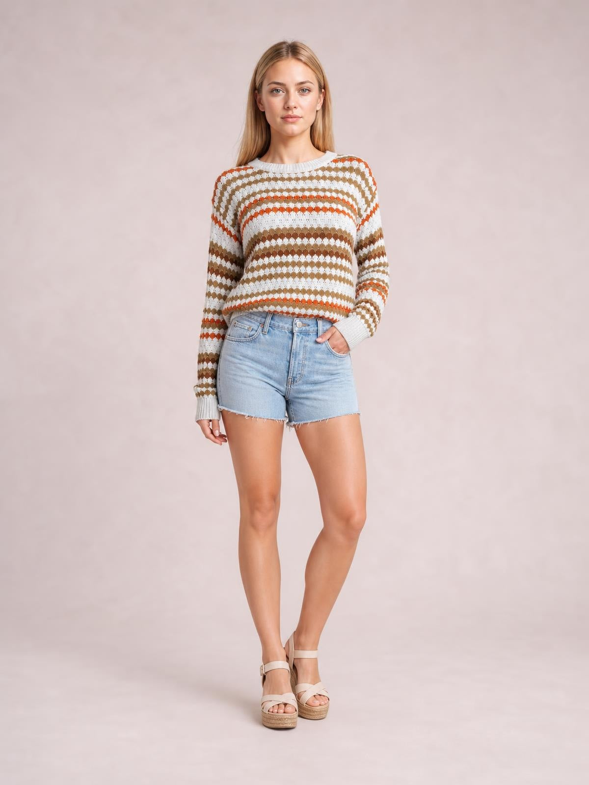 Model stands with one hand in her pocket wearing a cream drop-shoulder sweater with brown and orange striped eyelet pattern, ribbed trim, and relaxed silhouette.