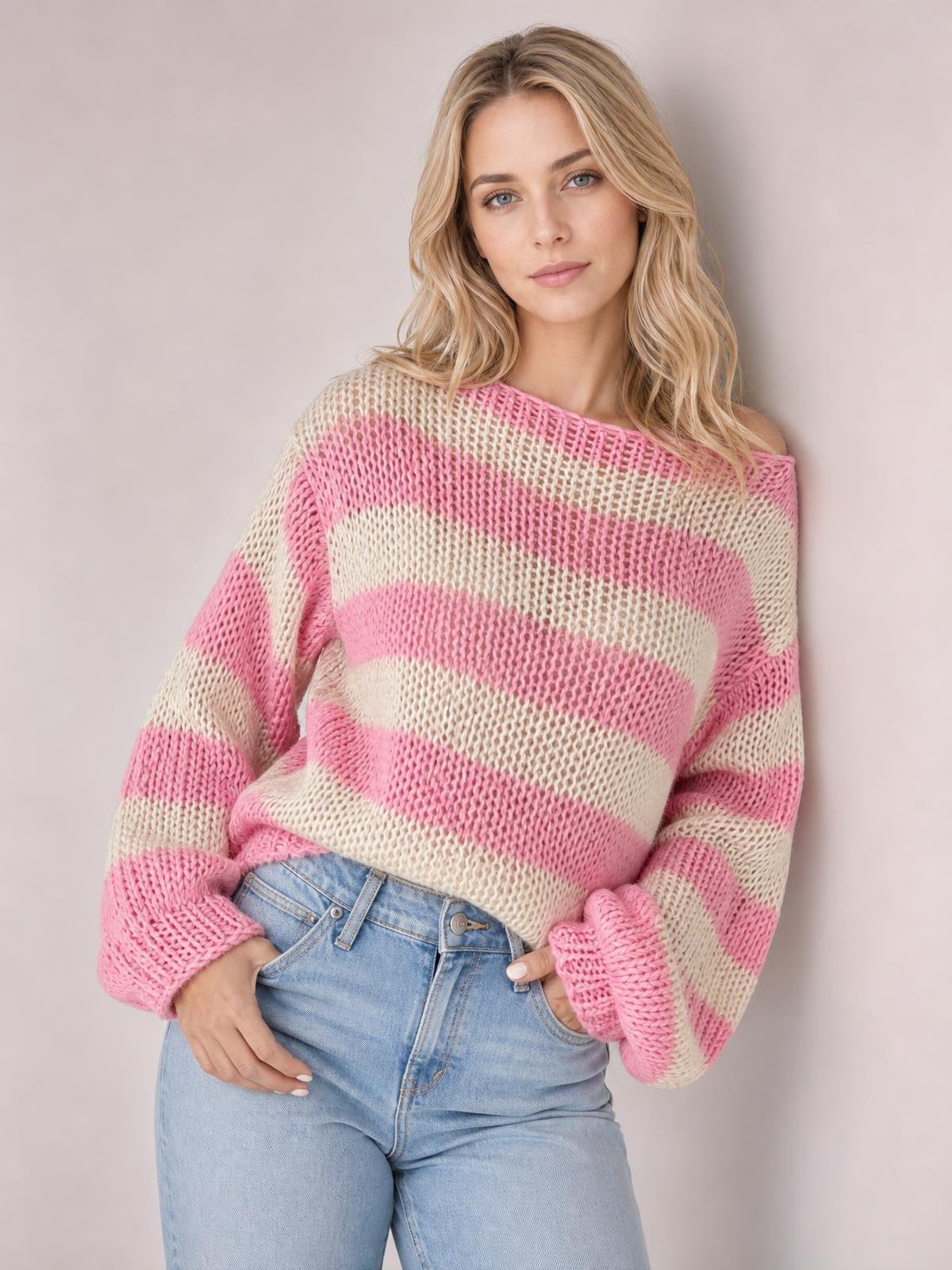 Model poses with hands at her jeans pockets in a pink and cream striped boat neck long sleeve sweater with a loose, open-knit silhouette.