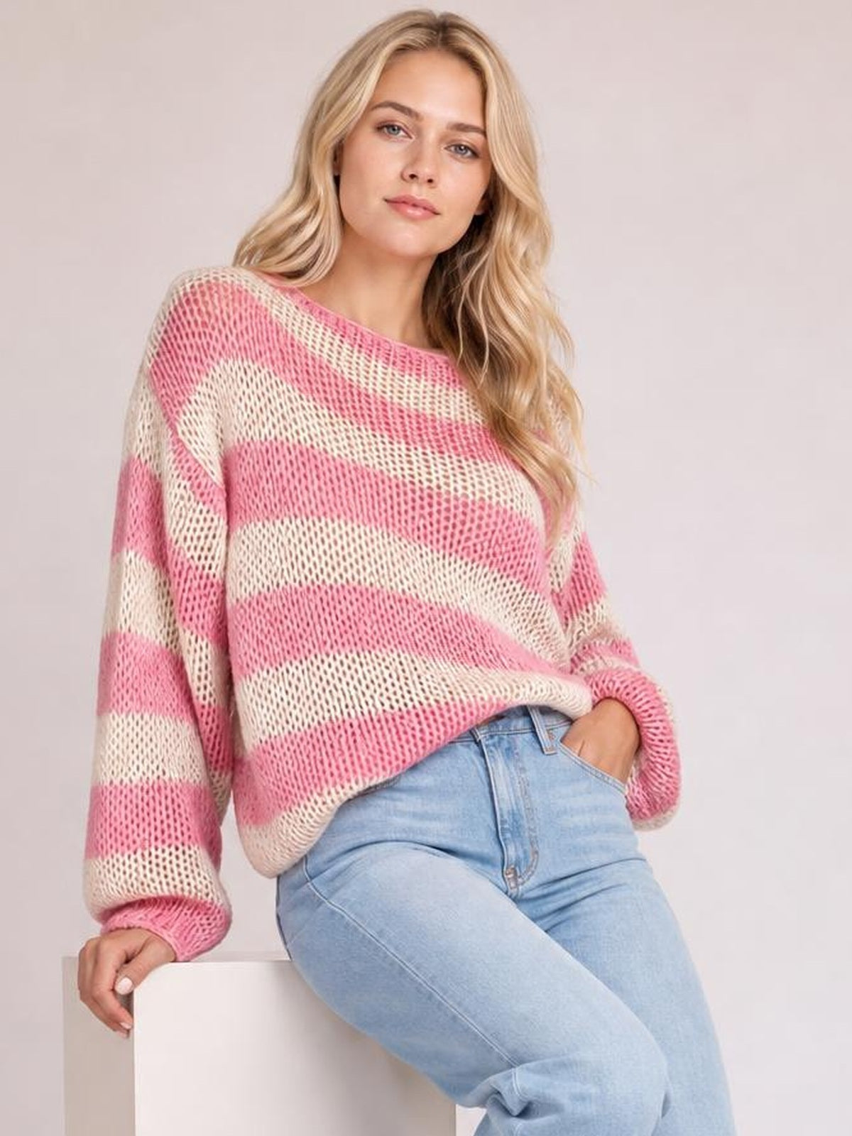 Model wearing a pink and cream striped boat neck long sleeve sweater with an open-knit texture and relaxed silhouette, leaning with one hand in her jeans pocket.