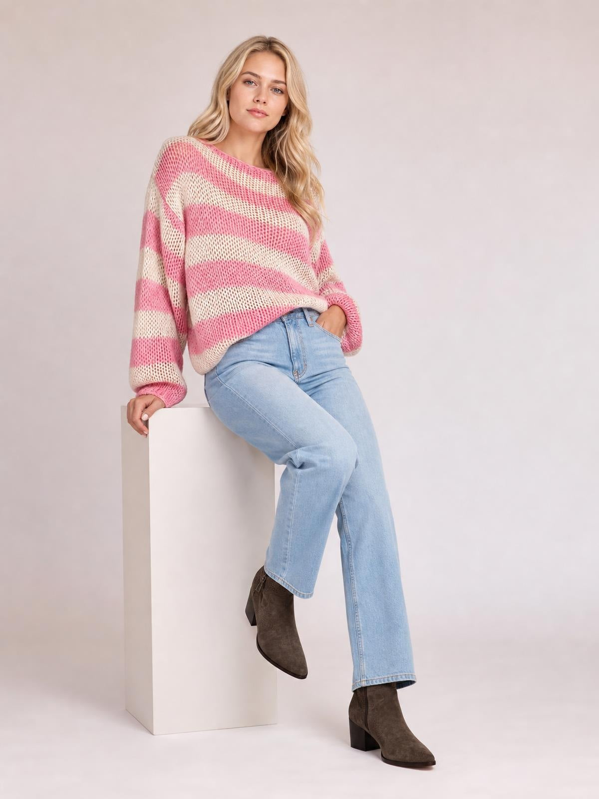 Model wears a pink and cream striped boat neck long sleeve sweater with a relaxed silhouette and dropped shoulders, posing with one hand in her jeans pocket.