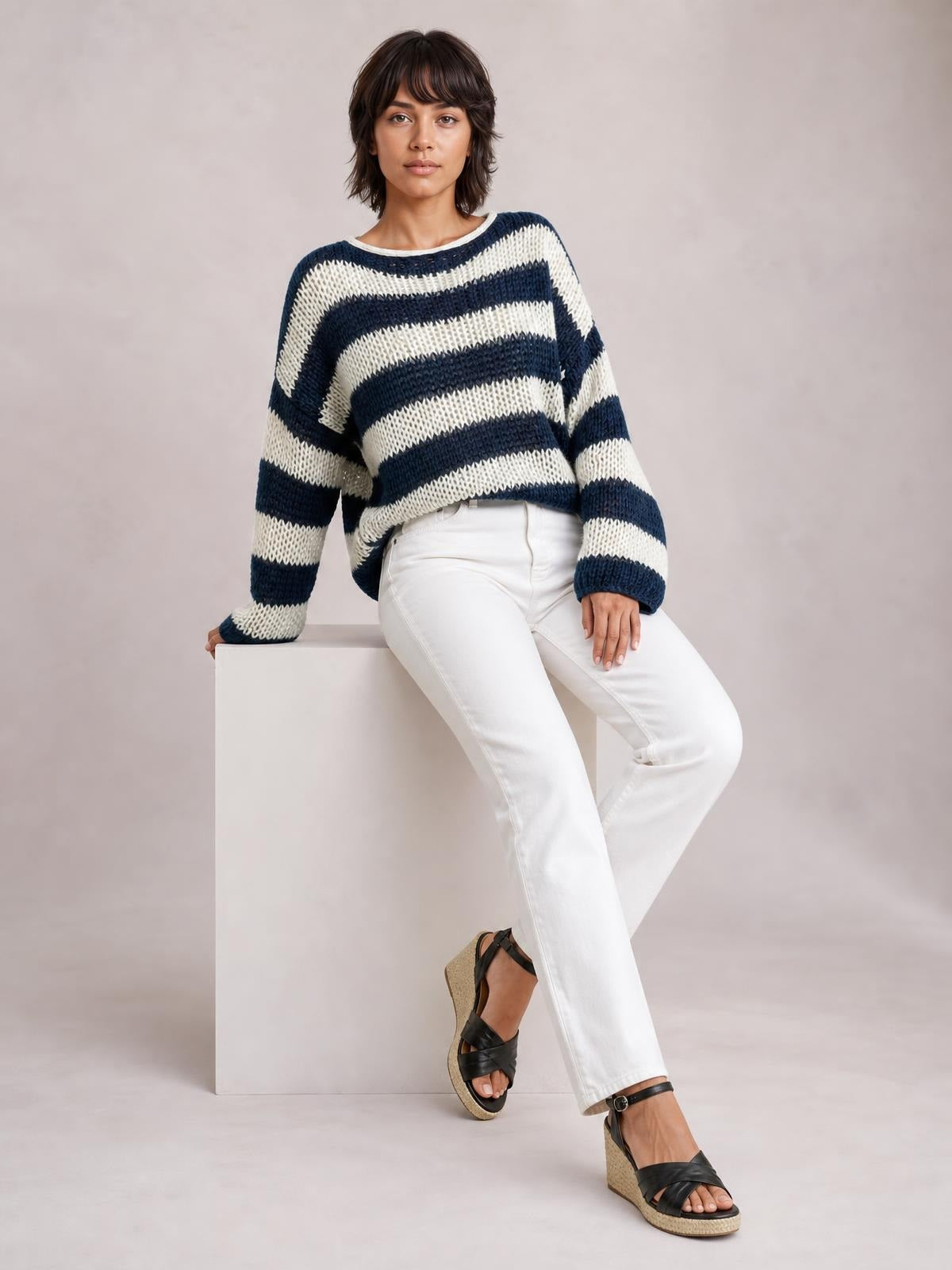Model seated in a relaxed pose wears a navy and white striped boat neck long sleeve sweater with a loose, slightly oversized silhouette and ribbed trim.