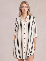 Model poses wearing a cream striped button-down shirt dress with vertical black stripes, a collared neckline, front buttons, rolled cuffs, and side pockets.